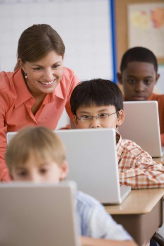 Female teacher helps student with laptop computer assignment as others students work on their laptops.